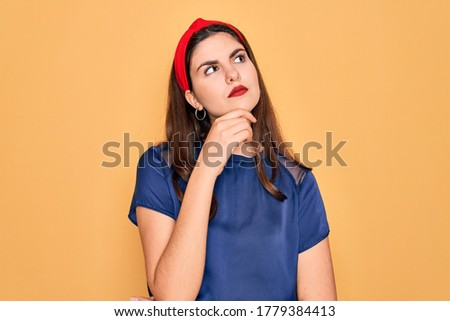Young beautiful brunette woman wearing red lips over yellow background with hand on chin thinking about question, pensive expression. Smiling with thoughtful face. Doubt concept.