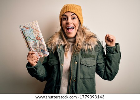 Young beautiful tourist woman wearing winter clothes holding city map over white background screaming proud and celebrating victory and success very excited, cheering emotion