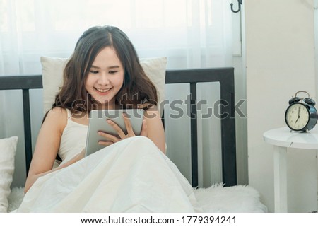 Beautiful asian woman just woke up and laying on the bed using tablet or video call in the morning light. Tablet , Gadget, Technology, Shopping concept. Copy Space.
