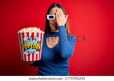 Young blonde woman wearing 3d glasses and eating pack of popcorn watching a movie on cinema covering one eye with hand, confident smile on face and surprise emotion.