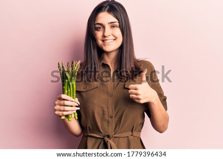 Young beautiful girl holding asparagus smiling happy and positive, thumb up doing excellent and approval sign 
