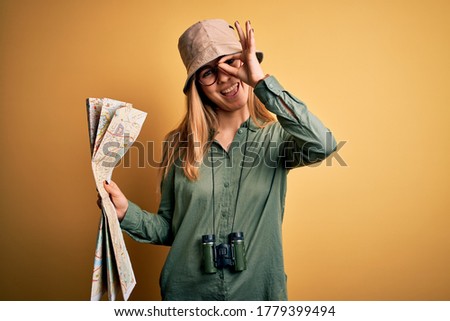 Blonde explorer woman with blue eyes on vacation wearing hat and binoculars holding city map with happy face smiling doing ok sign with hand on eye looking through fingers