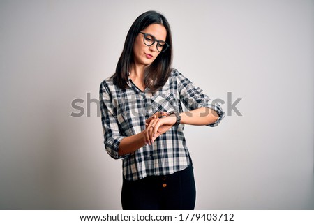 Young brunette woman with blue eyes wearing casual shirt and glasses over white background Checking the time on wrist watch, relaxed and confident