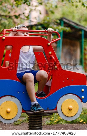 A boy in shorts and a T-shirt sits on a children's red car in the park in the summer. vertical photo