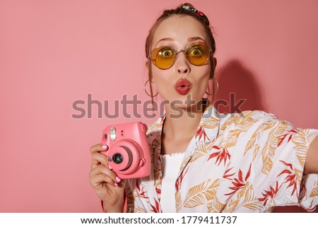Funny woman in stylish yellow sunglasses and round earrings in fashionable summer shirt making selfie and holding pink camera on isolated backdrop..