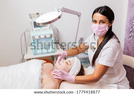 A cosmetologist in a medical mask and rubber gloves applies a cream mask to the client's face. Top view. In the background, a cosmetology device. Concept of cosmetology during the pandemic Royalty-Free Stock Photo #1779418877