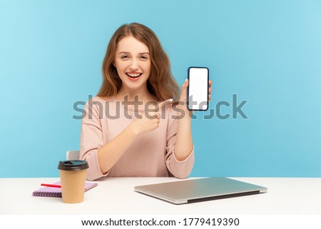 Joyful smiling woman, office employee, sitting at workplace and pointing mobile phone mock up display, copy space for application, website advertise. indoor studio shot isolated on blue background