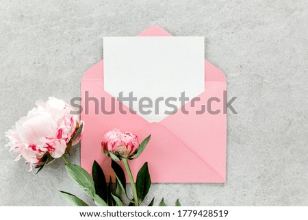 Mockup invitation, blank paper greeting card, pink envelope and peonies flower. Concrete table background. Flat lay, top view.