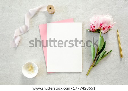 Mockup invitation, blank paper greeting card, pink envelope and peonies flower. Concrete table background. Flat lay, top view.