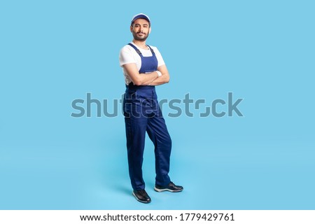 Full length confident professional handyman in overalls standing with crossed hands, smiling at camera. Profession of service industry, house repair. indoor studio shot isolated on blue background Royalty-Free Stock Photo #1779429761