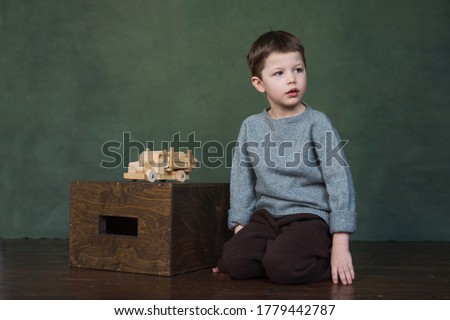handsome boy in knitted clothes sits on wooden floor, there is toy car on wooden box, good looks
