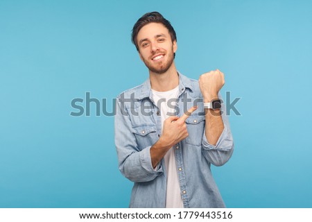 Please, hurry up! Portrait of cheerful punctual man in worker denim shirt pointing at wrist watch and smiling, showing smartwatch devise with mock up display. indoor isolated on blue background Royalty-Free Stock Photo #1779443516