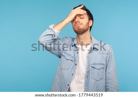 Portrait of upset man in worker denim shirt standing with facepalm gesture, blaming himself, feeling sorrow regret because of bad memory. studio shot isolated on blue background Royalty-Free Stock Photo #1779443519