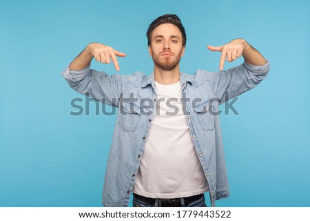 Here and right now! Portrait of bossy tyrant man in denim shirt pointing down and looking with arrogance, demanding immediate submission, demonstrating authority. indoor studio shot, blue background Royalty-Free Stock Photo #1779443522