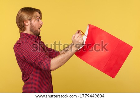 Scared surprised shopper guy in checkered shirt looking inside shopping bag with big eyes open mouth in amazement, shocked by purchase. indoor studio shot isolated on yellow background, side view