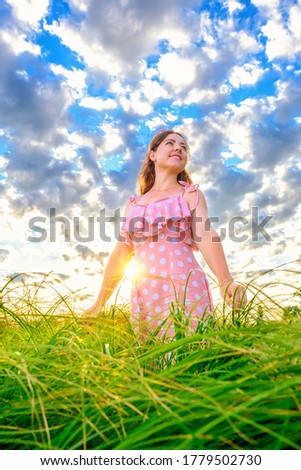 Young smiling caucasian girl standing with outstretched arms in grass on meadow against cloudy sky background 