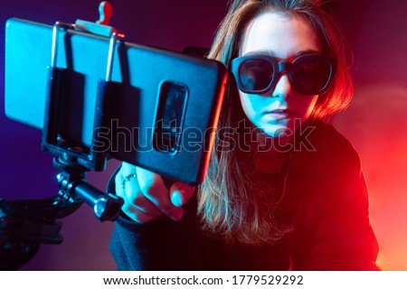 Girl video blogger. Phone mounted on a tripod in front of a woman. Girl student in dark. Young woman takes pictures of herself on phone. Video blogger is recording broadcast. Neon lighting.