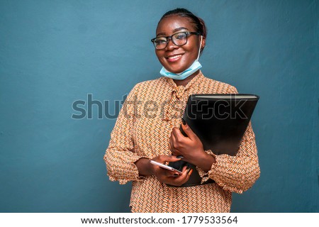 Beautiful African woman wearing spectacles, smiling, wearing surgical face mask for protection in covid-19 pandemic and holding a black case for documents and laptop,isolated on a blue background