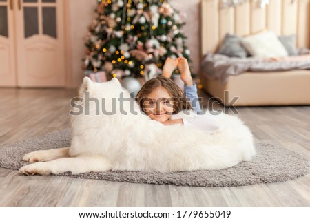 Christmas Child girl with smiling dog Samoyed lie on the floor in front of a large christmas tree. Christmas, winter and people concept. Christmas greeting card. Happy New Year. New Year at home