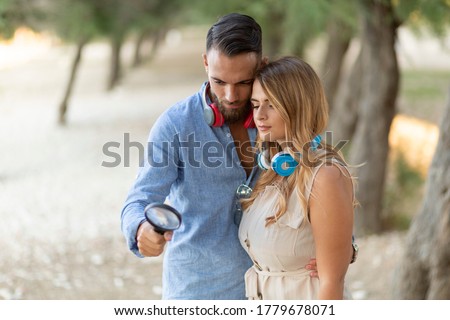 An Italian man with beard and red headphones and a magnifying glass and an Italian woman with cream dress and blue headphones with a magnifying glass looking for the right idea in the park among trees