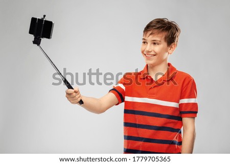 childhood, fashion and people concept - portrait of happy smiling boy in red polo t-shirt taking picture with smartphone on selfie stick over grey background