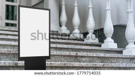 empty screen in black metal frame with white place for mock-up stands on steps of stone staircase near entrance to house or hotel outside