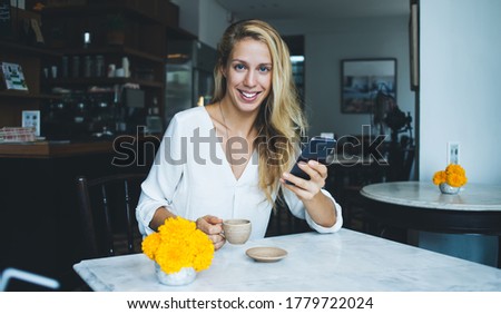 Gorgeous joyful female in casual clothes looking at camera and smiling while using smartphone at table with bright yellow flowers having aromatic hot drink in contemporary coffee shop