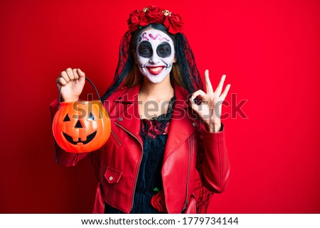 Woman wearing day of the dead costume holding pumpkin doing ok sign with fingers, smiling friendly gesturing excellent symbol 