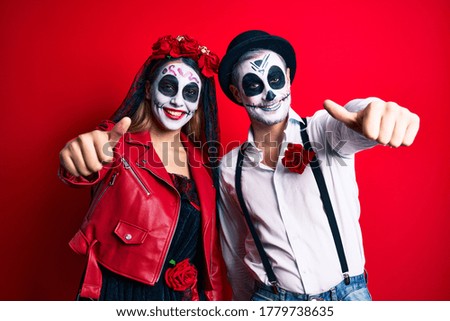Couple wearing day of the dead costume over red approving doing positive gesture with hand, thumbs up smiling and happy for success. winner gesture. 