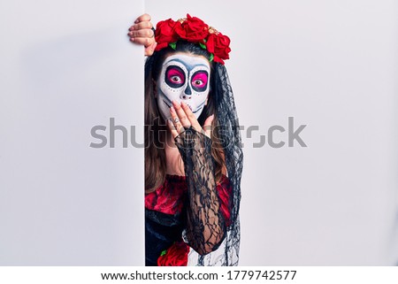 Young woman wearing day of the dead custome holding blank empty banner shocked covering mouth with hands for mistake. secret concept. 