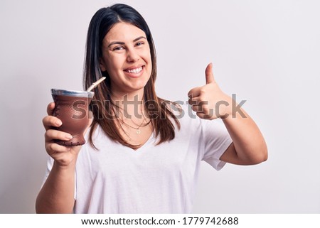Young beautiful brunette woman drinking mate infusion beverage over white background smiling happy and positive, thumb up doing excellent and approval sign