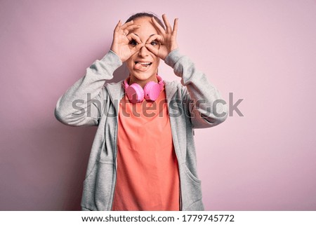 Beautiful sporty woman doing sport listening to music using headphones over pink background doing ok gesture like binoculars sticking tongue out, eyes looking through fingers. Crazy expression.