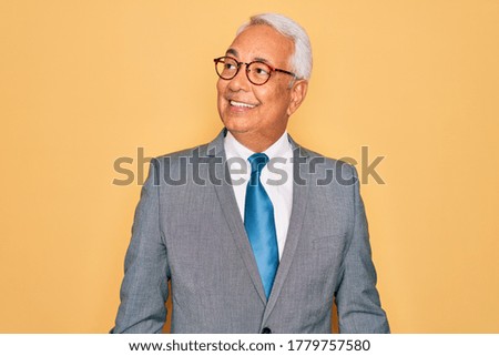 Middle age senior grey-haired handsome business man wearing glasses over yellow background looking away to side with smile on face, natural expression. Laughing confident.