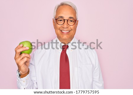 Middle age senior grey-haired nutritionist man holding healthy green apple over pink background with a happy face standing and smiling with a confident smile showing teeth