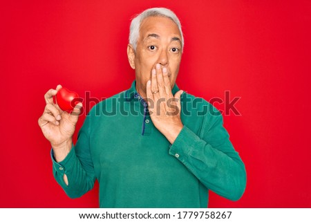 Middle age senior grey-haired man holding romantic red heart object over isolated red background cover mouth with hand shocked with shame for mistake, expression of fear, scared in silence, secret