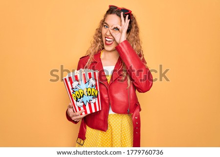 Beautiful blonde woman with blue eyes eating salty popcorn snack over yellow background with happy face smiling doing ok sign with hand on eye looking through fingers