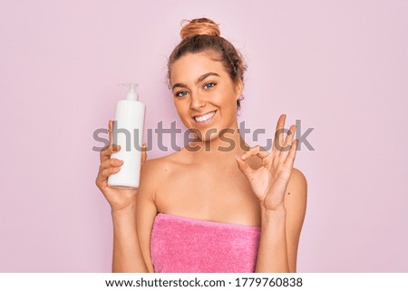 Beautiful woman with blue eyes wearing towel shower after bath holding bottle of body cream doing ok sign with fingers, excellent symbol