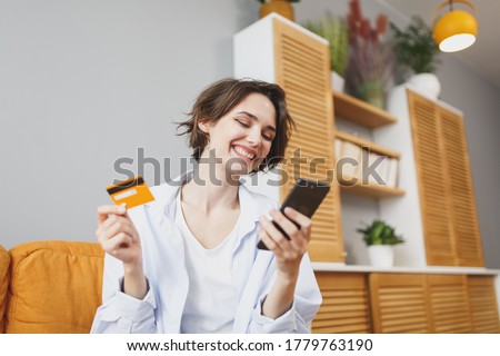 Smiling young woman in casual clothes sit on couch spending time in living room at home. Rest relax good mood leisure lifestyle concept. Mock up copy space. Using mobile phone, hold credit bank card