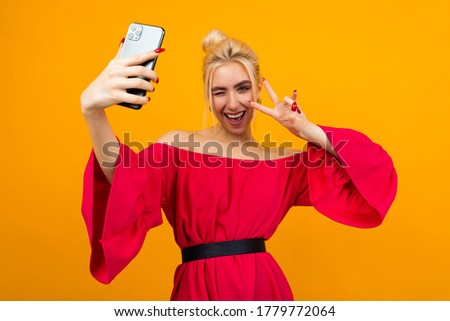 girl in an elegant red dress makes a photo of herself on the phone on a yellow studio background