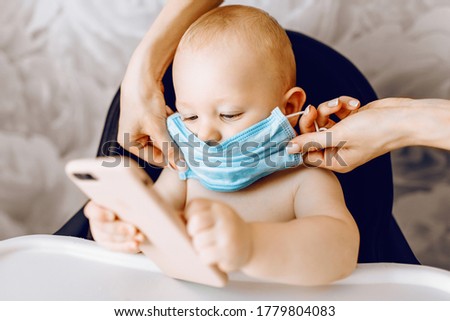 small child, wearing a medical mask on his face, is sitting in a feeding chair with a mobile phone, watching cartoons. Quarantine, coronavirus