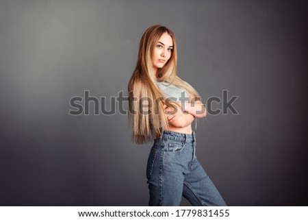 Photo portrait of a beautiful woman girl crossed her arms on her chest on a gray background in jeans with long blonde beautiful hair. Standing in front of a camera with a serious face