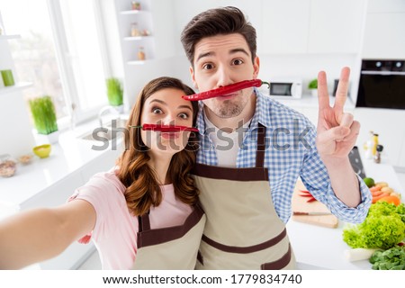 Two people dream comic humor joking couple lovers have fake mustache red hot chilli pepper make selfie show v-sign enjoy healthy weekend lifestyle prepare nutrition in kitchen house indoors