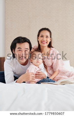 Young happy Vietnamese husband and wife hugging little son when watching cartoon or movie on tv set