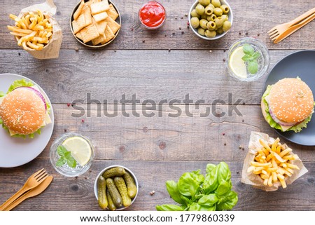 Dinner table with burger, french fries, vegetables, sauces, snacks and lemonade on wooden background, top view. Social distance after a pandemic, copy space.