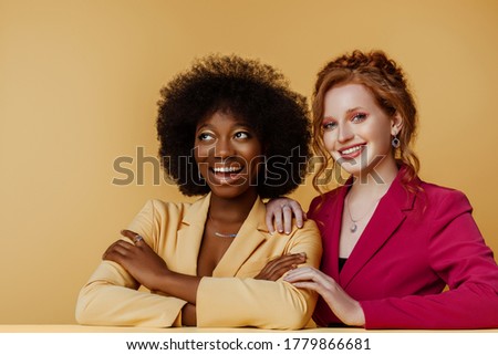 Diverse beauty, fashion: portrait of two happy smiling multiethnic women wearing colorful blazers, posing on yellow background. Different nation, multiracial friends concept. Copy space for text