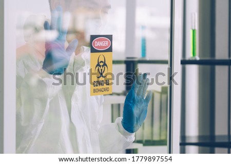 Lab Technician in Personal protective equipment  PPE suit touching windows glass from inside of Laboratory with biohazard sign