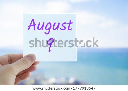 August 9th. Hand holding sticker with text August 9 on the blurred background of the sea and sky. Copy space for text. Month in calendar concept