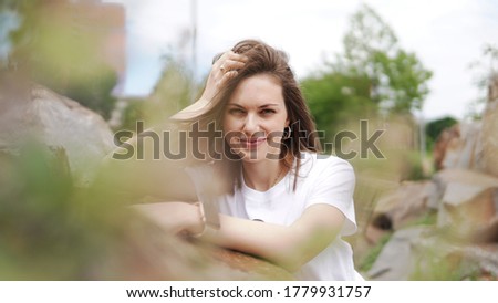 Beautiful woman in white shirt near artificial rocks in park. Young smiling woman with long hair putting hands on stones