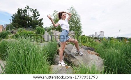 Beautiful woman in white shirt on artificial rocks in park. Young woman with long hair standing with raised hands on stones