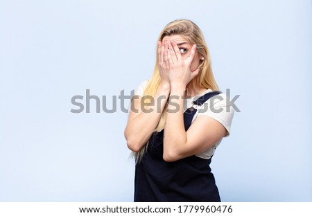young pretty blonde woman covering face with hands, peeking between fingers with surprised expression and looking to the side against flat wall
